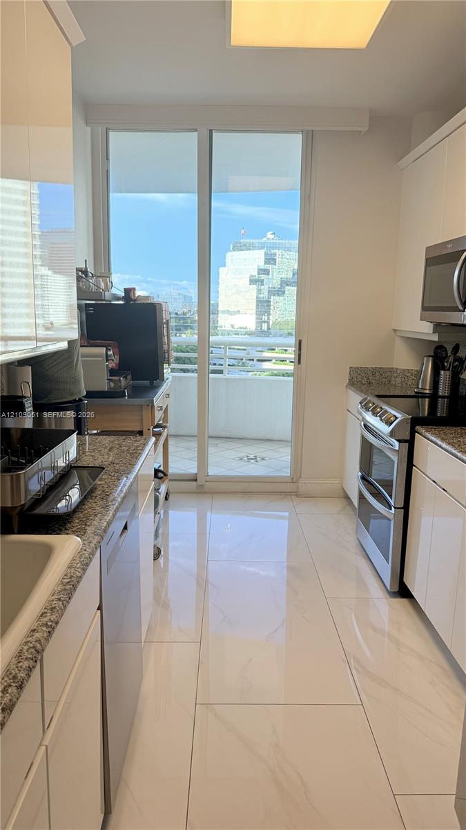 Kitchen featuring high-gloss white cabinetry, stone-finish countertops, stainless steel appliances, large-format tile flooring, and a glass sliding door opening to an exterior balcony