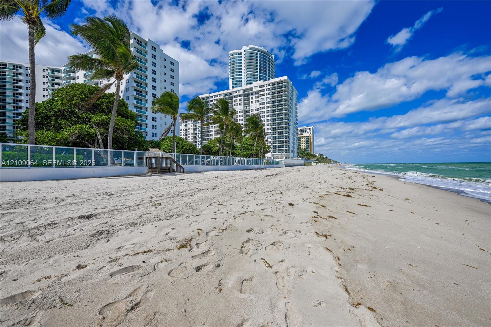 Sandy beach shoreline featuring direct coastal access, glass-panel perimeter fencing, and mature palm trees