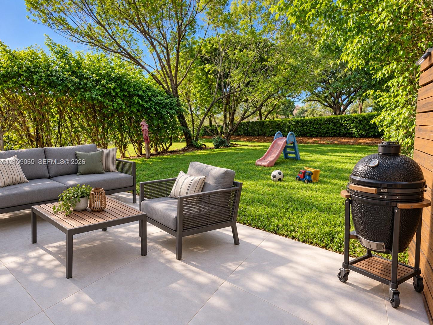 Rear patio featuring large-format light-toned stone tile flooring, horizontal wood-slat privacy fencing, and a grassy lawn bordered by mature trees and manicured hedges