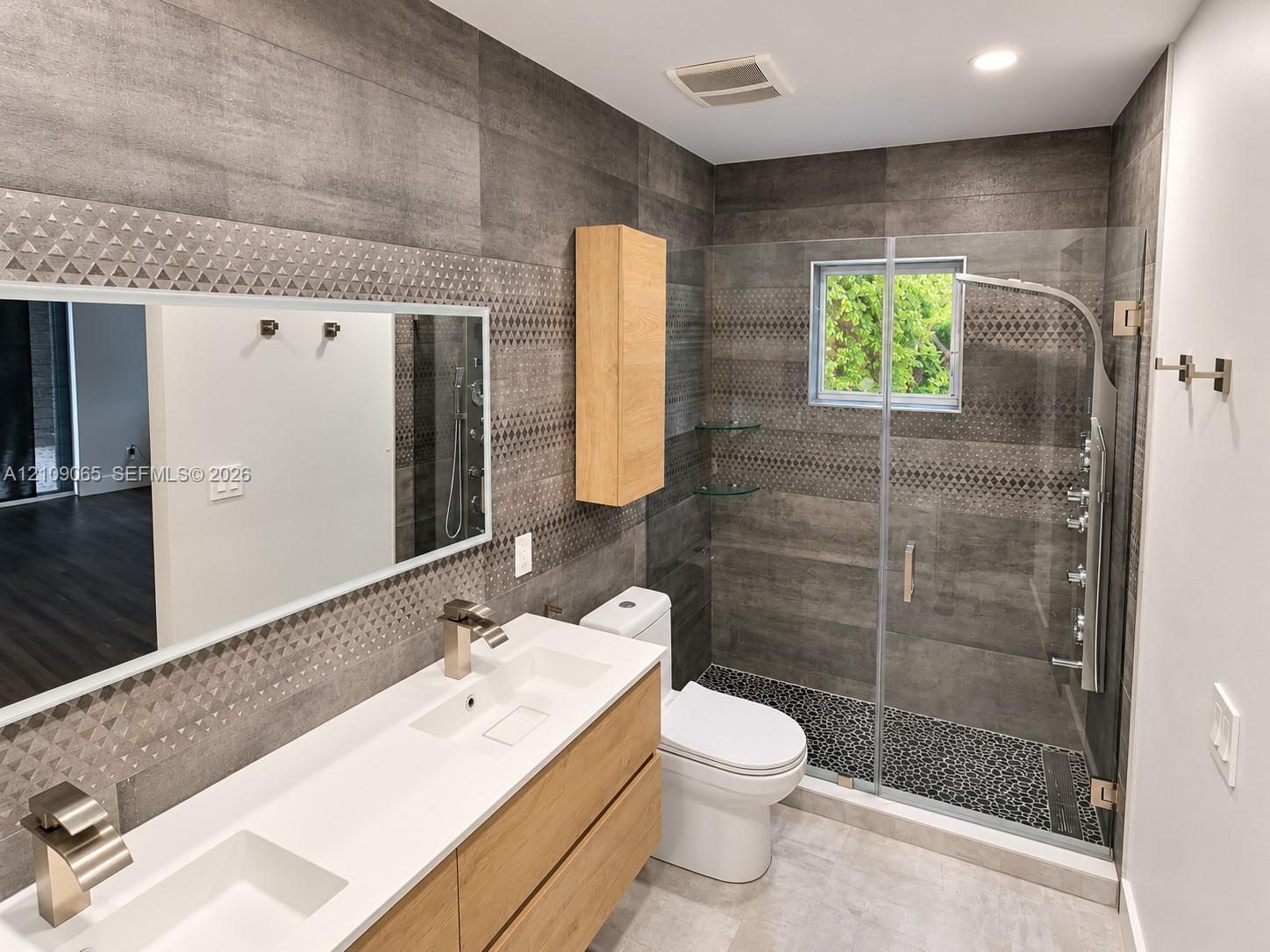 Bathroom featuring a dual-sink vanity with a white countertop, large-format grey wall tile with a geometric accent band, glass-enclosed shower with body spray fixtures and stone-tiled floor, and wood-finish cabinetry