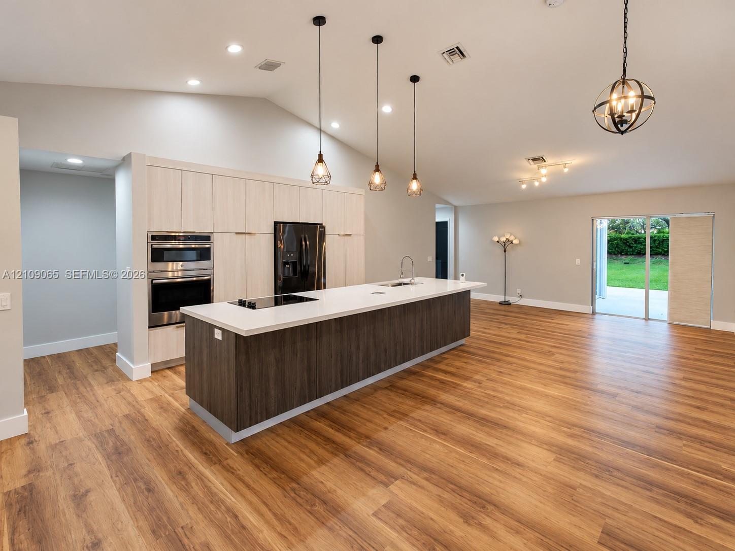 Open-concept kitchen featuring vaulted ceilings, wood-finish flooring, a central island with a solid surface countertop, light-toned cabinetry, and built-in double wall ovens