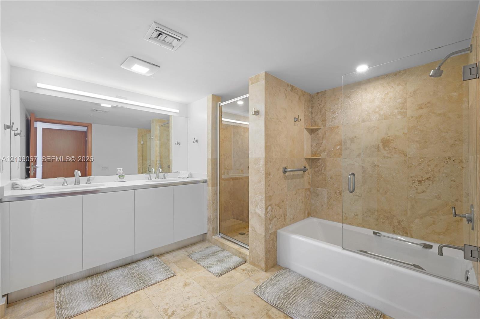 Double vanity with rectangular sinks, expansive wall-to-wall mirror, tiled soaking tub with glass partition, and separate walk-in shower featuring natural stone wall tile Double vanity with rectangular sinks, expansive wall-to-wall mirror, tiled soaking tub with glass partition, and separate walk-in shower featuring natural stone wall tile