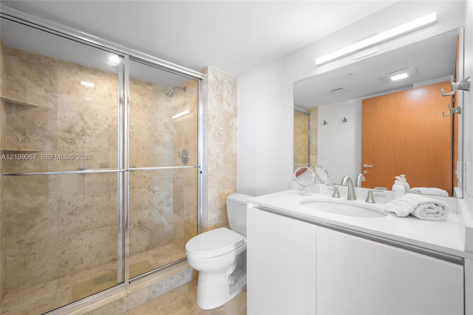 Walk-in shower with stone tile surround, glass sliding door, white vanity with integrated basin, polished metal hardware, and large vanity mirror