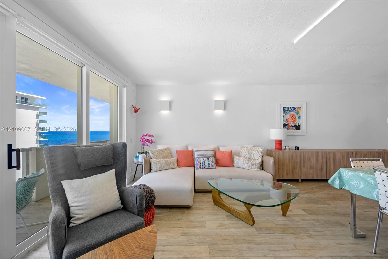 Living area featuring wood-finish flooring, floor-to-ceiling sliding glass doors, wall-mounted light fixtures, and expansive ocean and coastal scenery Living area featuring wood-finish flooring, floor-to-ceiling sliding glass doors, wall-mounted light fixtures, and expansive ocean and coastal scenery