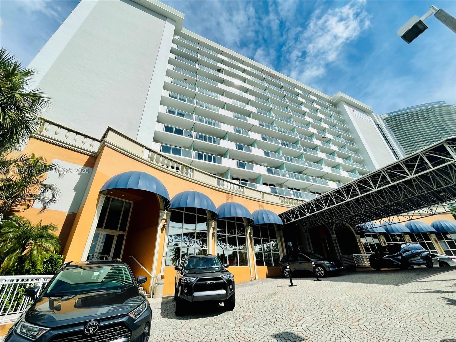 Multi-story high-rise exterior featuring a paved arrival court, decorative metal porte-cochre, arched window awnings, and tiered balconies with glass railings