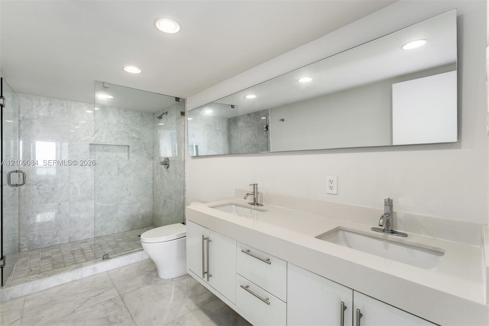 Dual-sink vanity with quartz countertop, walk-in glass-enclosed shower featuring stone-finish tiling, full-width vanity mirror, and recessed lighting