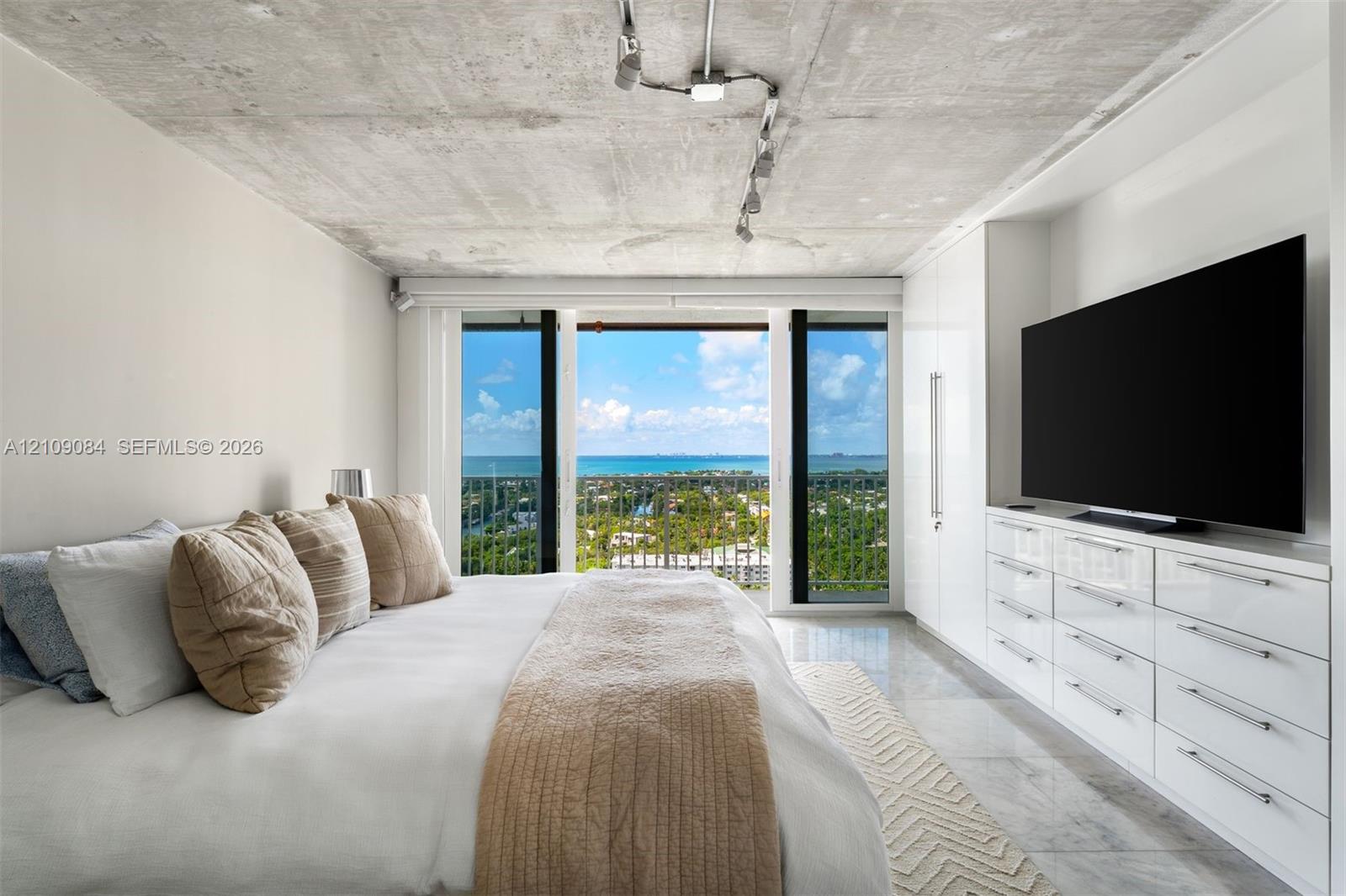 Exposed concrete ceiling with industrial track lighting, floor-to-ceiling glass paneling, built-in high-gloss white cabinetry, polished tile flooring, and panoramic ocean backdrop