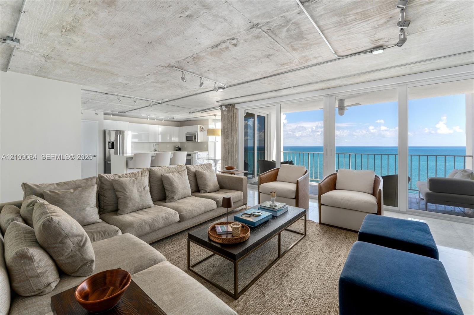 Open-concept living space featuring exposed concrete ceilings, polished stone flooring, custom wall-mounted cabinetry, track lighting, and floor-to-ceiling glass sliding doors with ocean scenery