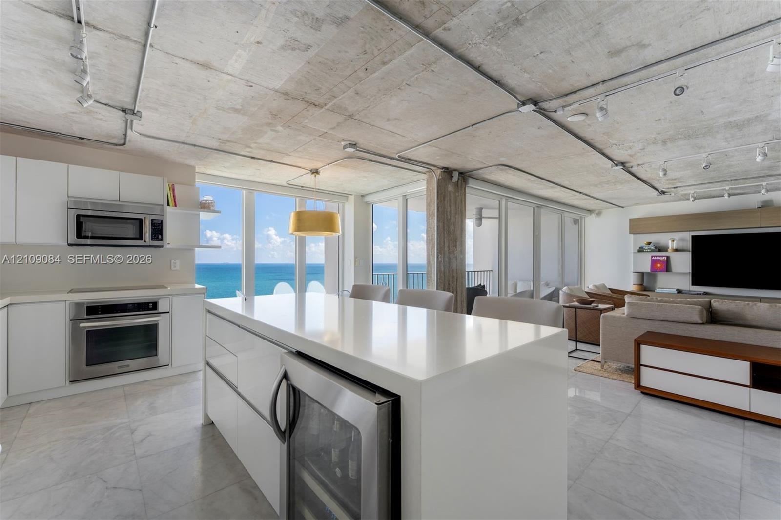 Exposed concrete ceiling with industrial-style track lighting, expansive floor-to-ceiling glass sliding doors, private balcony, and ocean backdrop