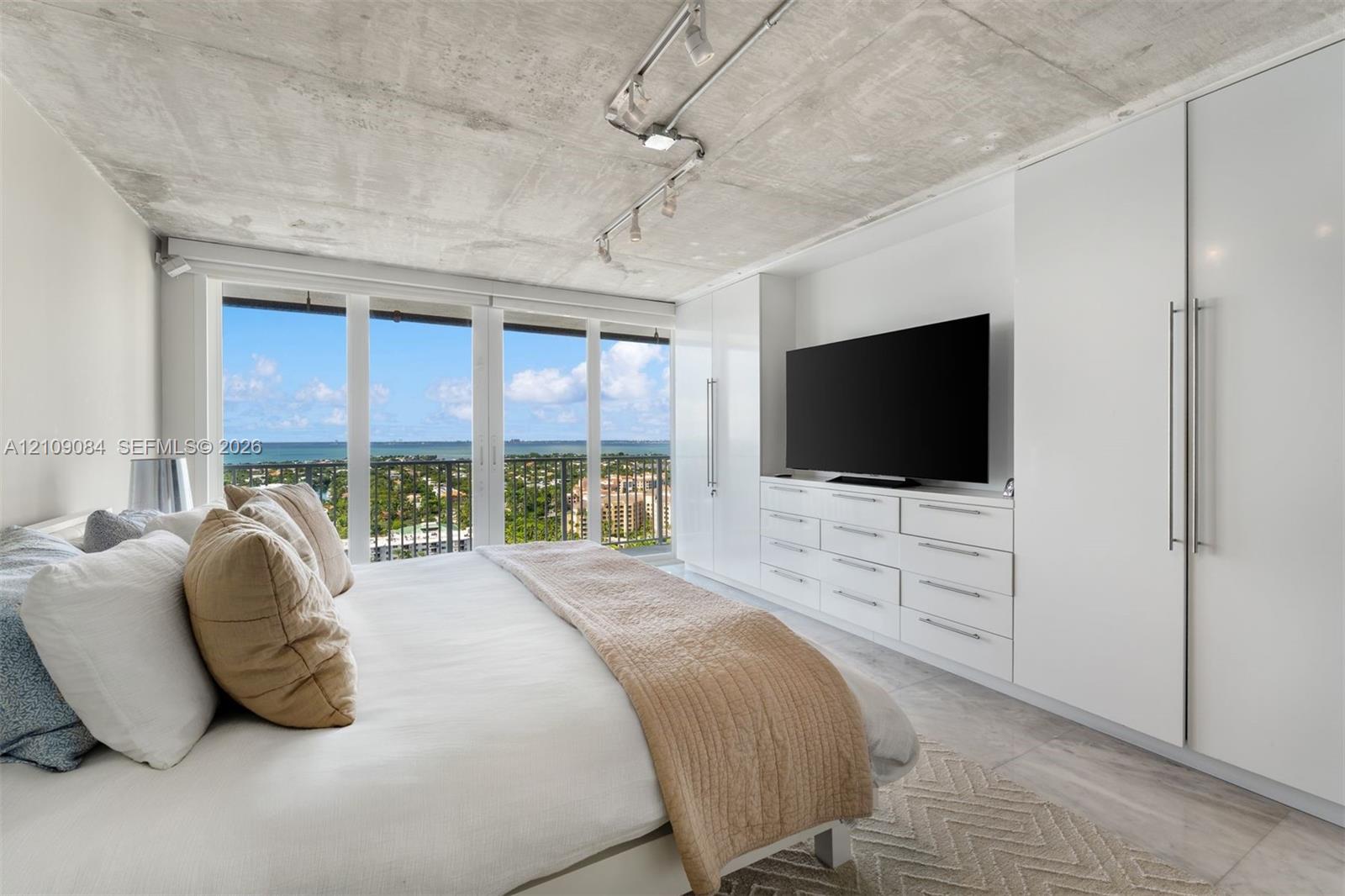 Exposed concrete ceiling with mounted track lighting, floor-to-ceiling windows, custom built-in white cabinetry, and large-format tile flooring