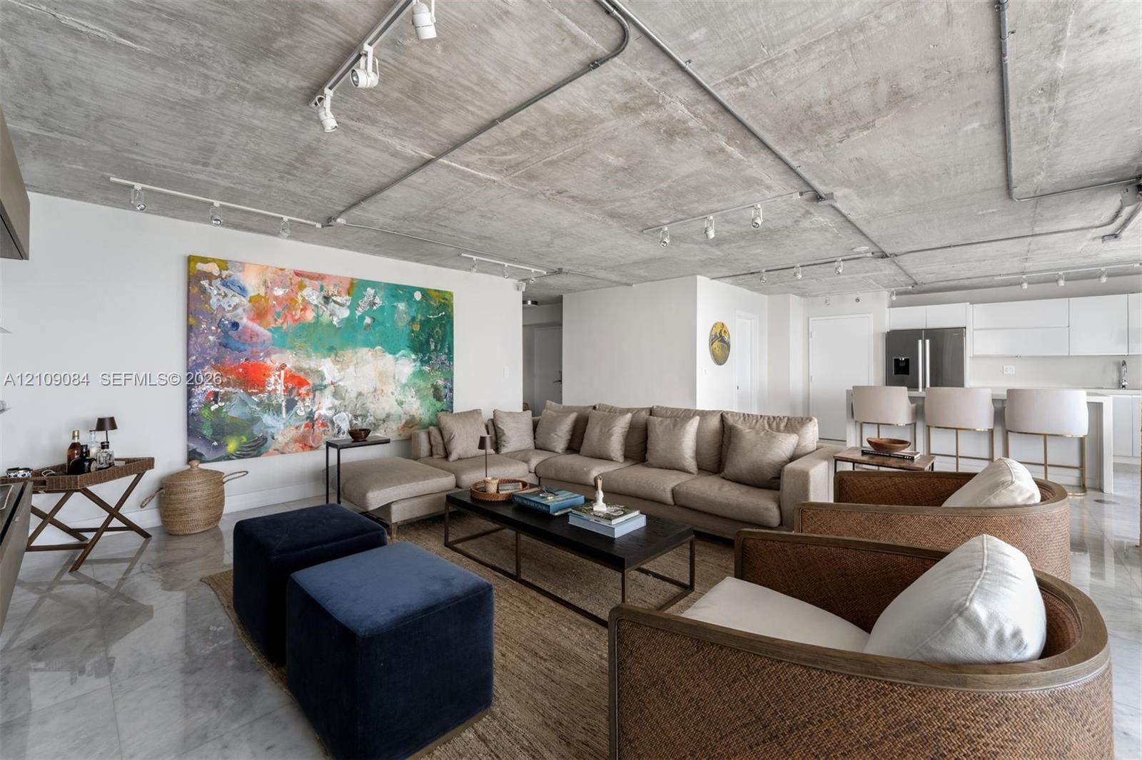 Exposed concrete ceiling with industrial track lighting, floor-to-ceiling glass doors, wall-mounted light wood-finish cabinetry, and balcony access featuring ocean water scenery