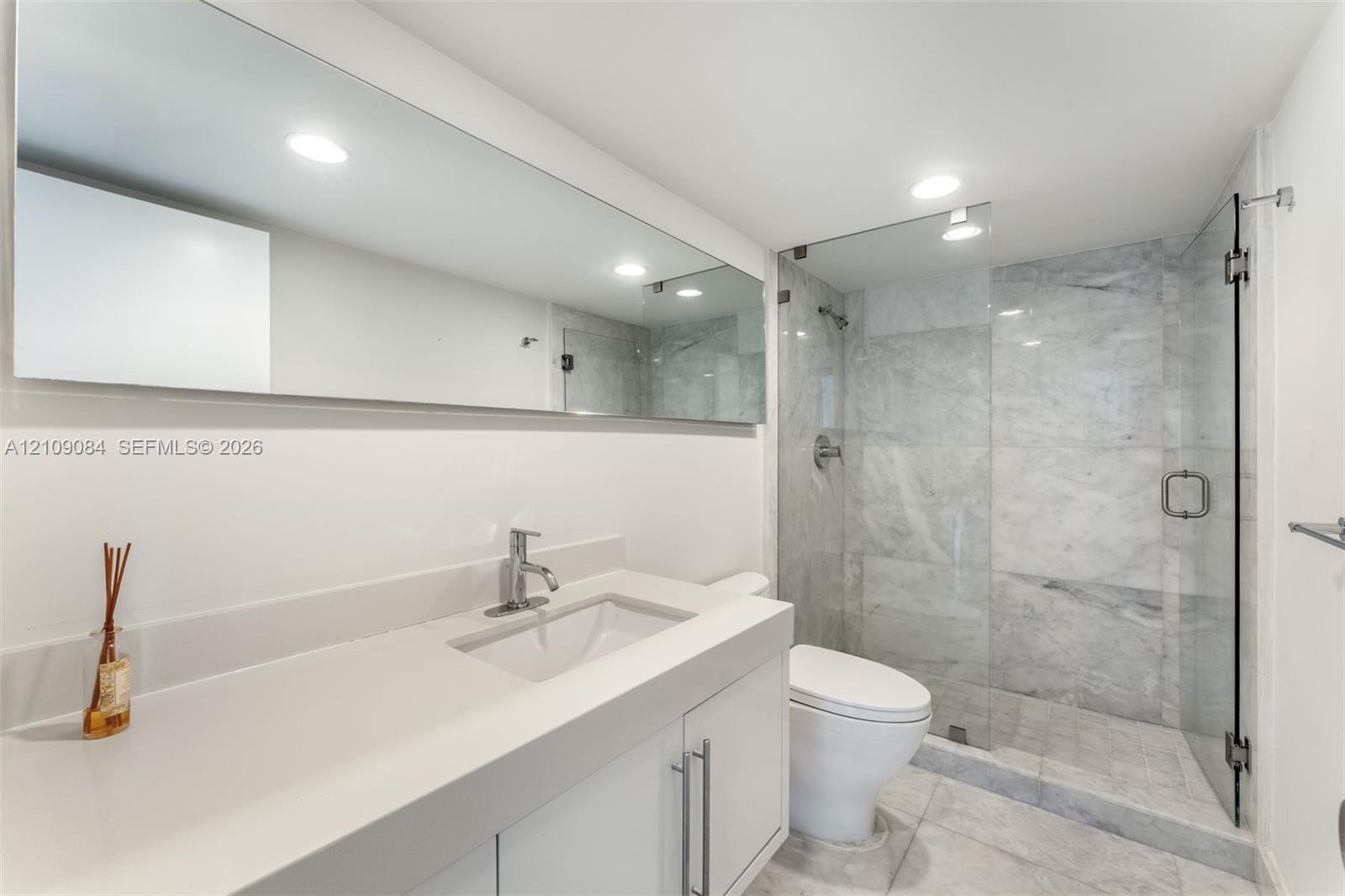 White vanity with a solid-surface countertop and integrated basin, full-width mirror, and walk-in shower featuring glass paneling and stone-tile surrounds