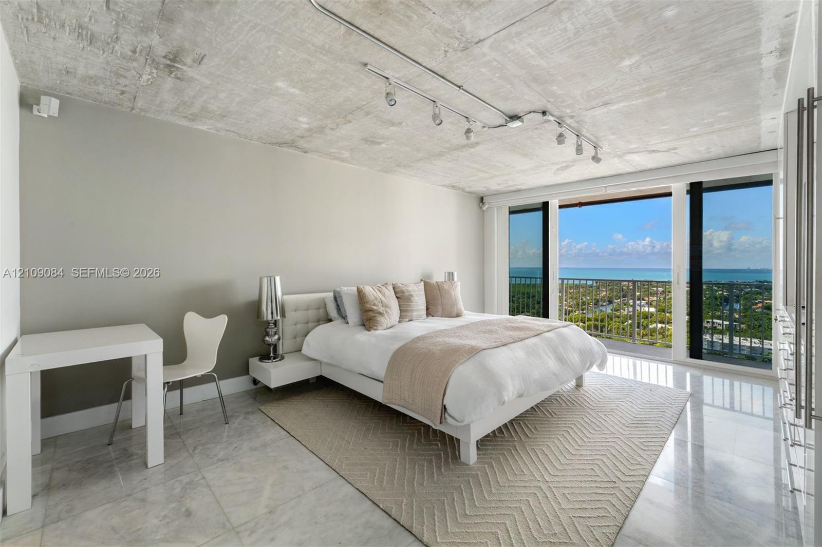 Exposed concrete ceiling, track lighting, floor-to-ceiling glass doors, large format stone flooring, and metal railing balcony