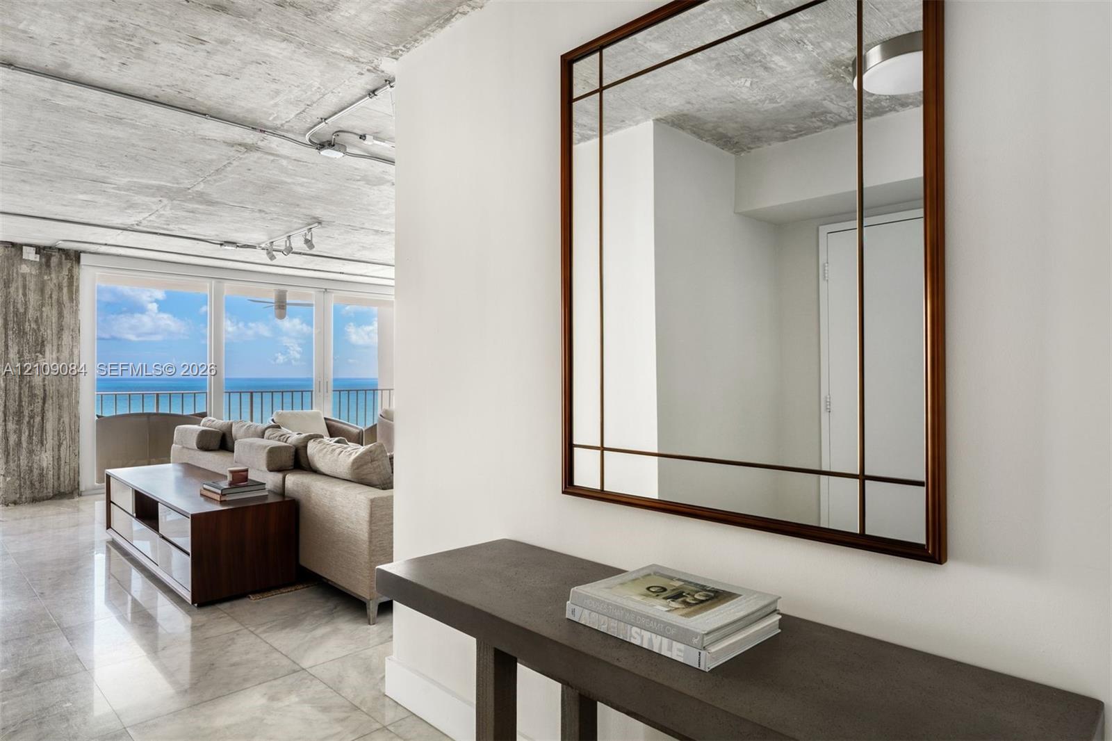 Exposed concrete ceilings, large-format tile flooring, wall-mounted track lighting, and full-height glass doors showcasing ocean scenery