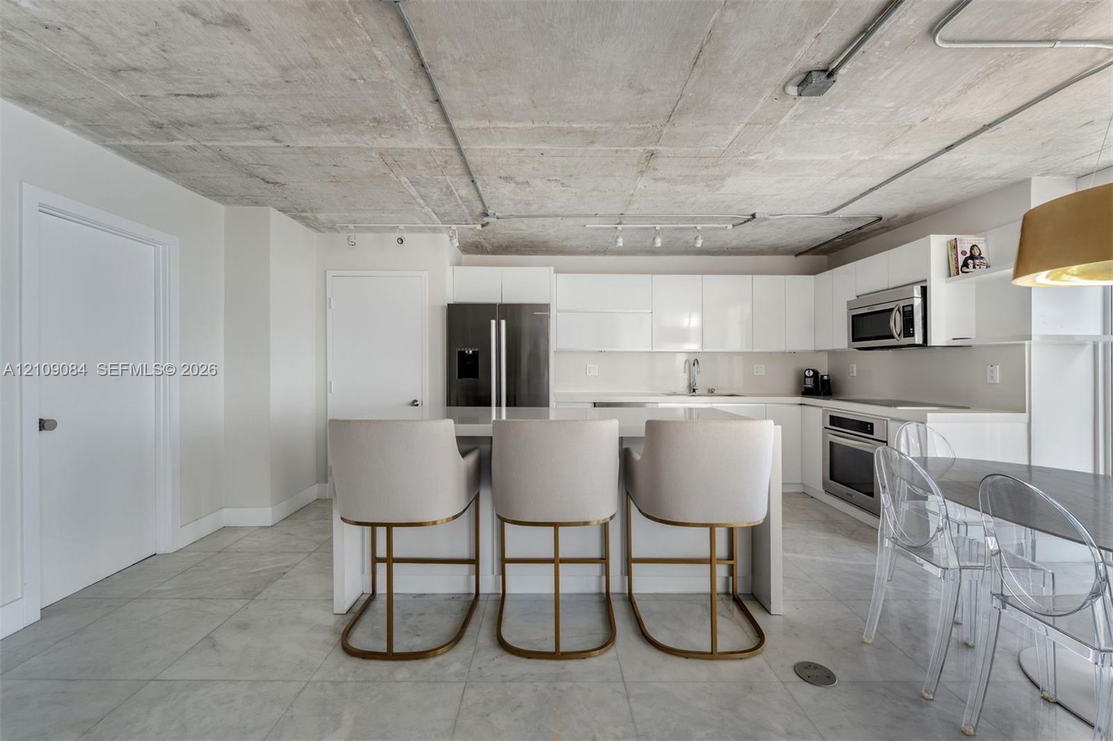 Modern kitchen featuring a central island with a quartz countertop, exposed concrete ceiling with conduit lighting, flat-panel white cabinetry, stainless steel appliances, and polished stone flooring