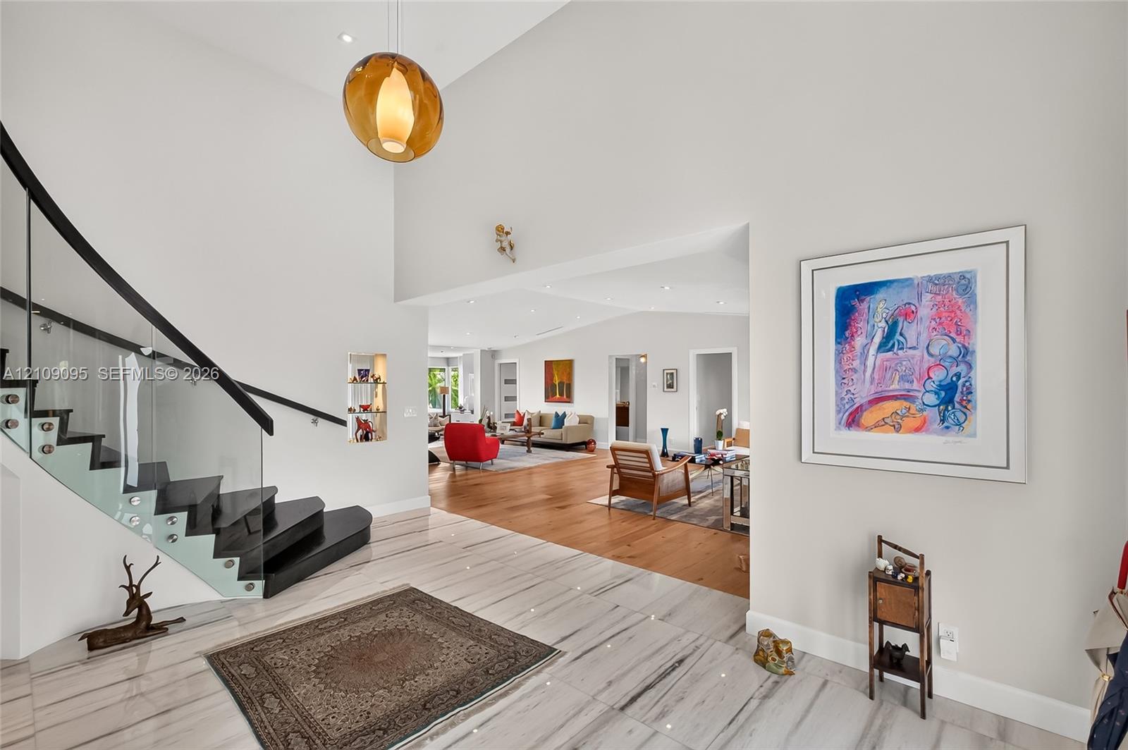 Double-height ceiling, floating staircase with glass paneling and dark-finish treads, stone-style tile flooring, wood-finish flooring, and statement pendant lighting