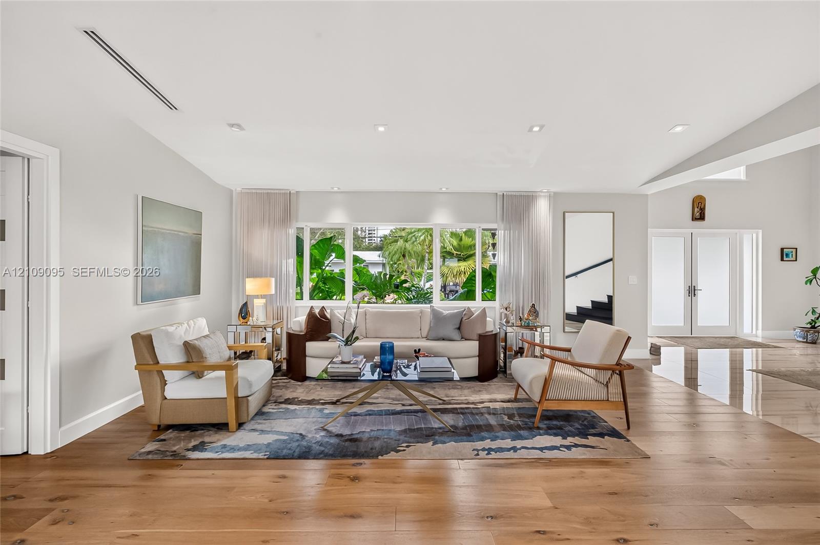 Open-concept living space featuring wood-finish flooring, recessed lighting, double-entry doors, and large windows with exterior greenery