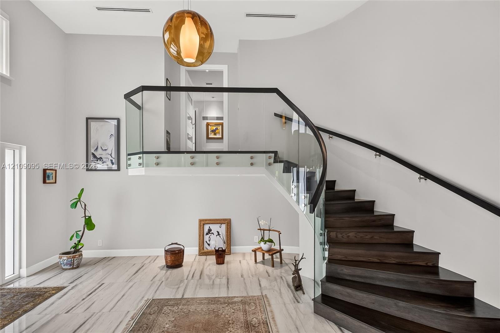 Curved staircase featuring dark-finished wood treads, glass panel railings, and a minimal handrail, accented by a pendant light fixture and polished stone-finish flooring