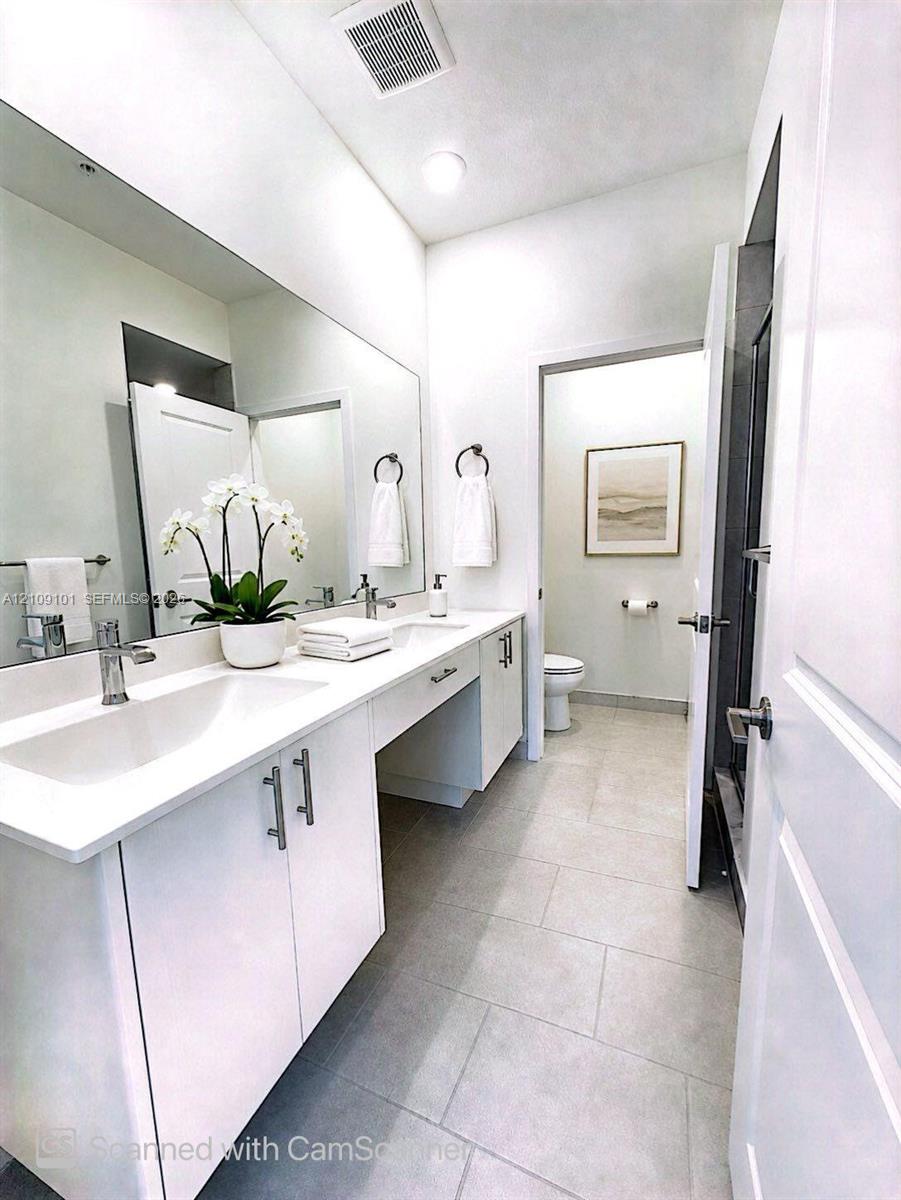 Dual-sink vanity with white cabinetry, sleek hardware, expansive wall-to-wall mirror, and neutral tile flooring
