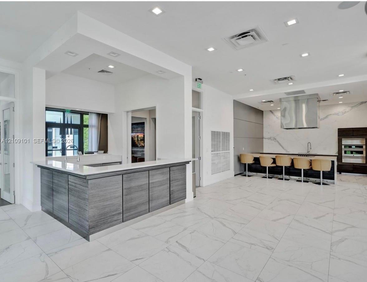 Kitchen featuring large-format tile flooring, a dual-level island with stone countertops and wood-finish cabinetry, a full-height stone backsplash, and stainless steel hood range