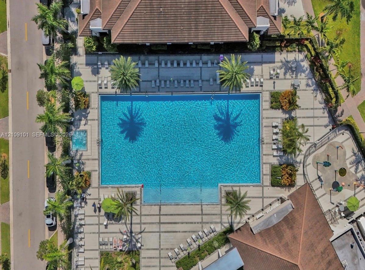 Aerial perspective featuring a large swimming pool with an integrated spa, expansive stone-paver pool deck, structured landscaping with mature palm trees, and a splash pad area