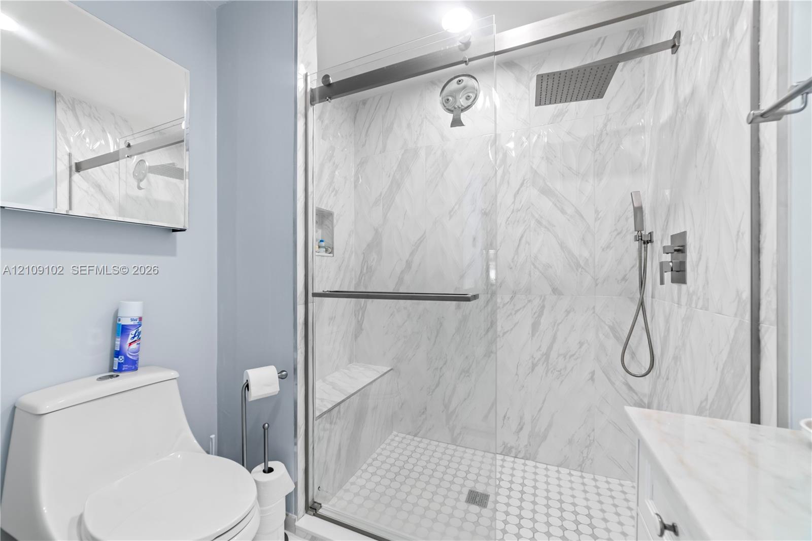 Walk-in shower featuring floor-to-ceiling stone-finish tile, glass sliding enclosure, built-in bench, rain-style shower head, and handheld sprayer