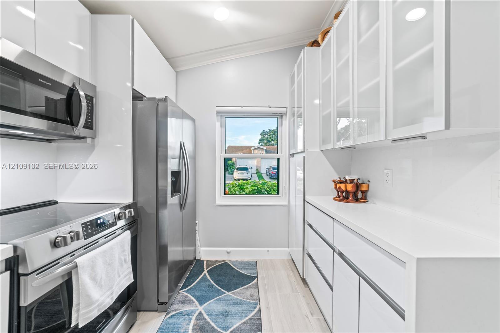 Kitchen featuring high-gloss white cabinetry, white countertop surfaces, stainless steel appliances, glass-front upper cabinets, and light-toned flooring