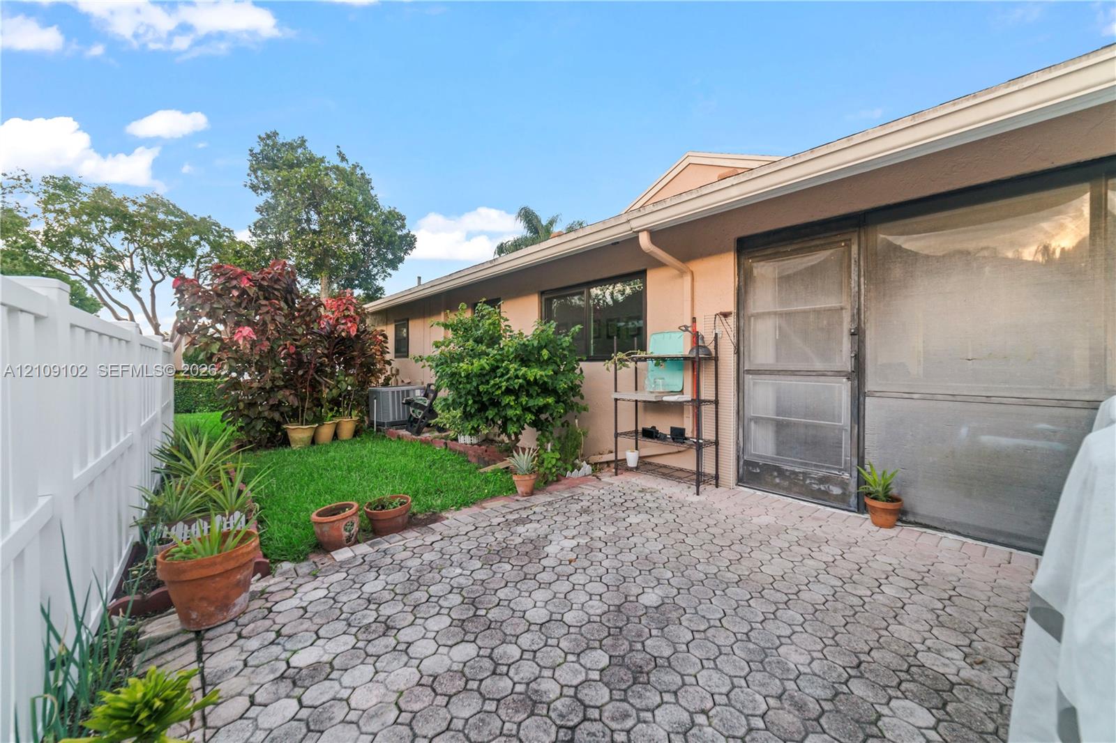 Exterior patio featuring interlocking concrete pavers, enclosed by a white vinyl privacy fence, with a screened-in entrance and landscaped green space