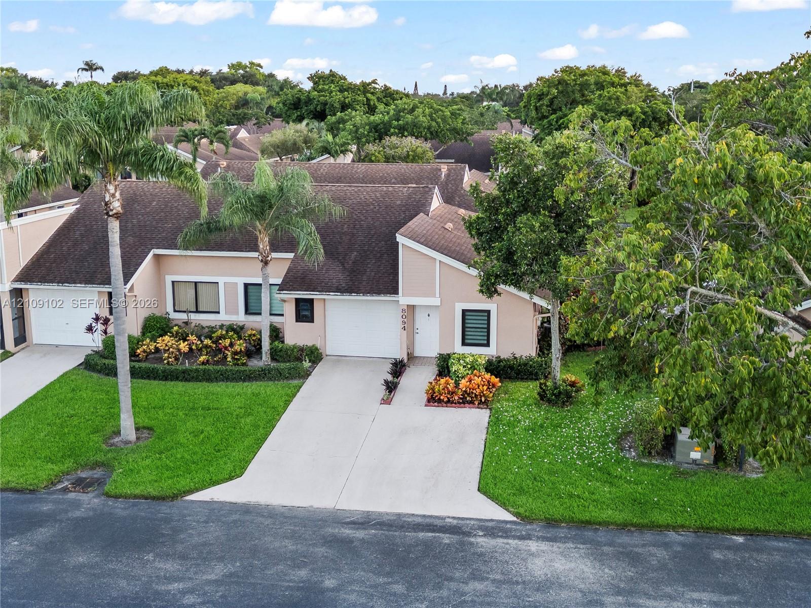 Single-story exterior featuring a shingled roof, concrete driveway, two-car garage, and established tropical landscaping