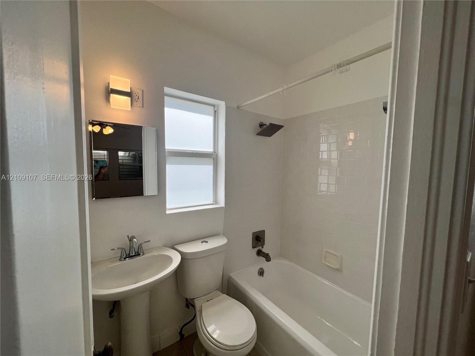 Bathroom featuring a pedestal sink, wall-mounted rectangular vanity light, bathtub with subway tile surround, and matte-finish shower fixtures