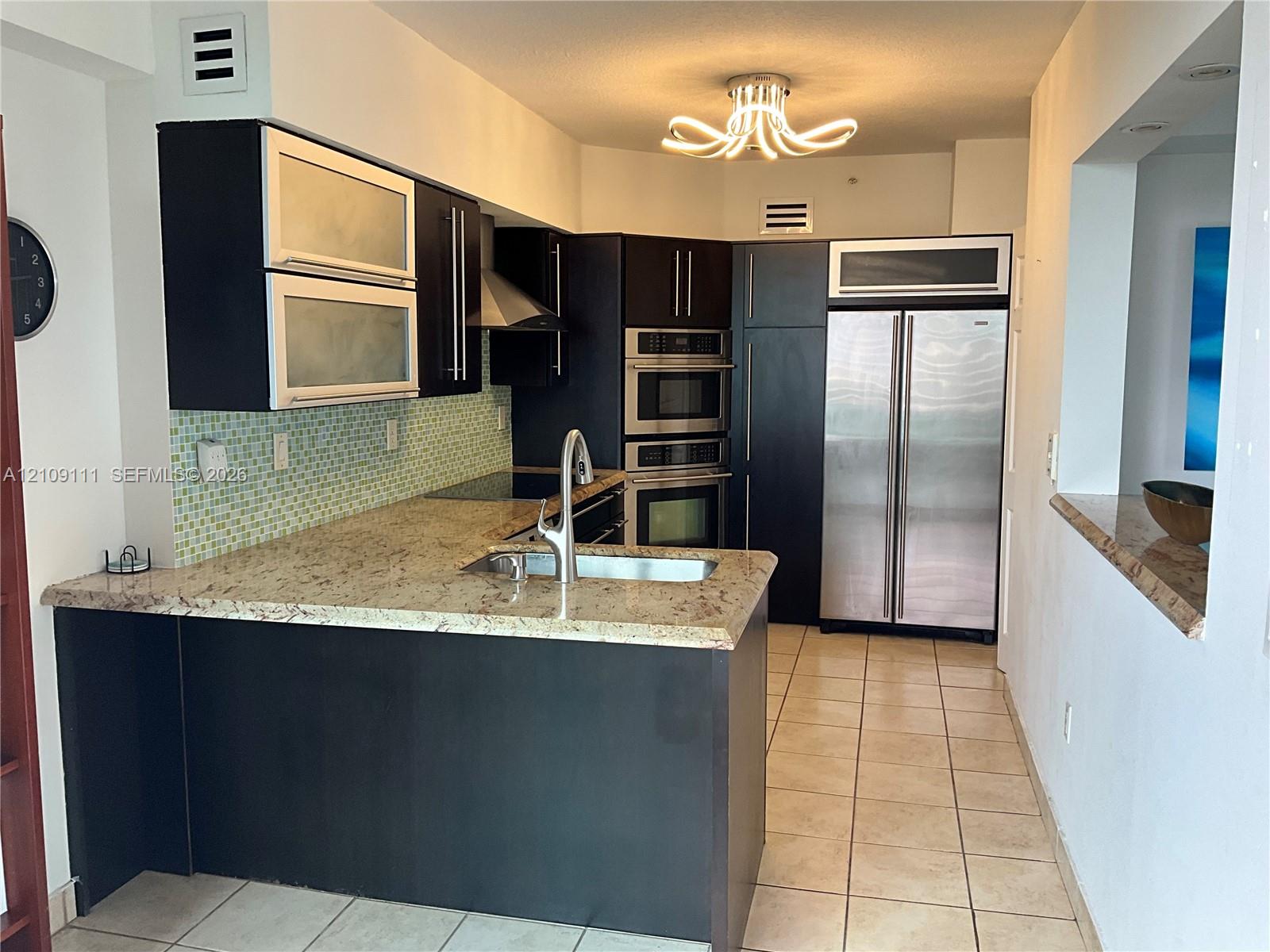 Kitchen featuring dark cabinetry, stone countertops, a mosaic tile backsplash, built-in stainless steel appliances, and a peninsula with a stainless steel sink