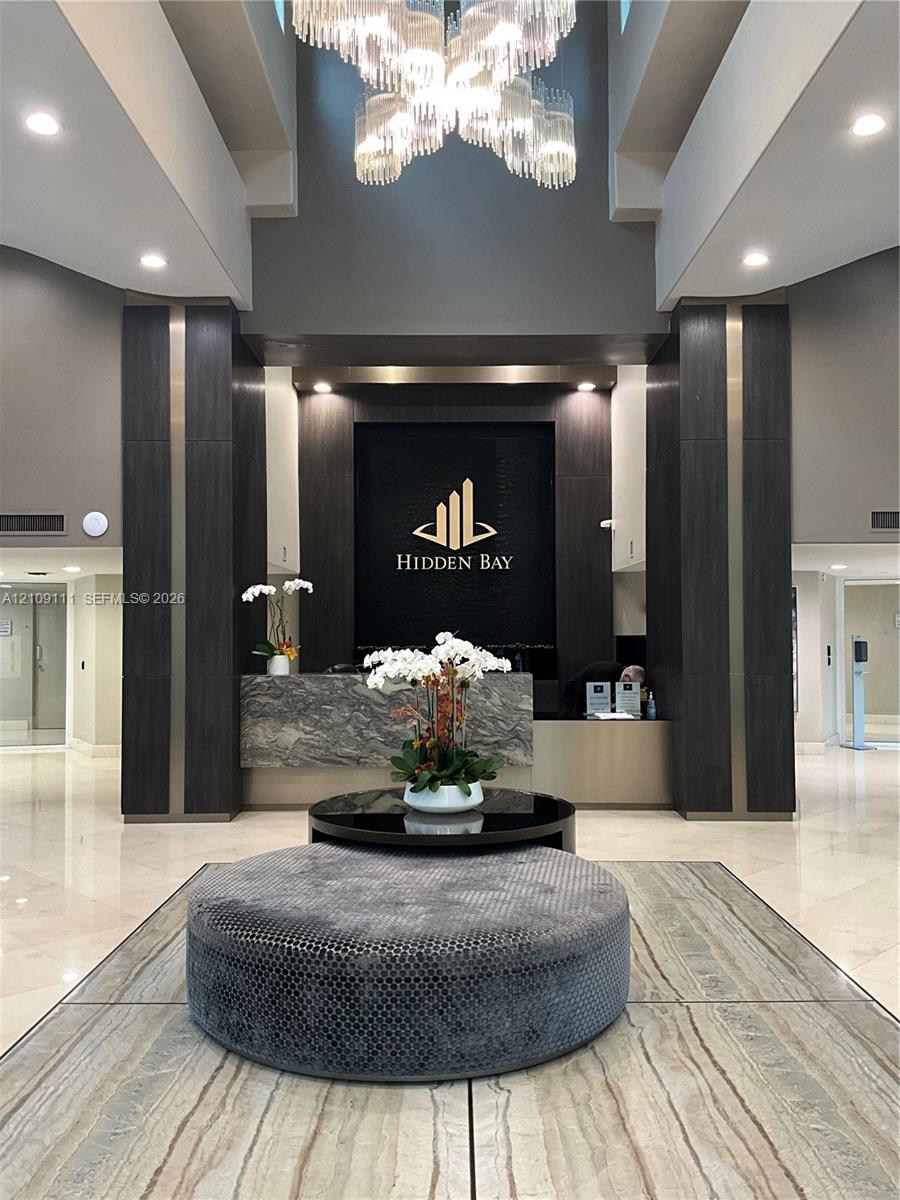 Grand lobby featuring a tiered crystal chandelier, stone-finish reception desk, recessed lighting, and polished stone flooring with vertical wood-paneled accents Grand lobby featuring a tiered crystal chandelier, stone-finish reception desk, recessed lighting, and polished stone flooring with vertical wood-paneled accents