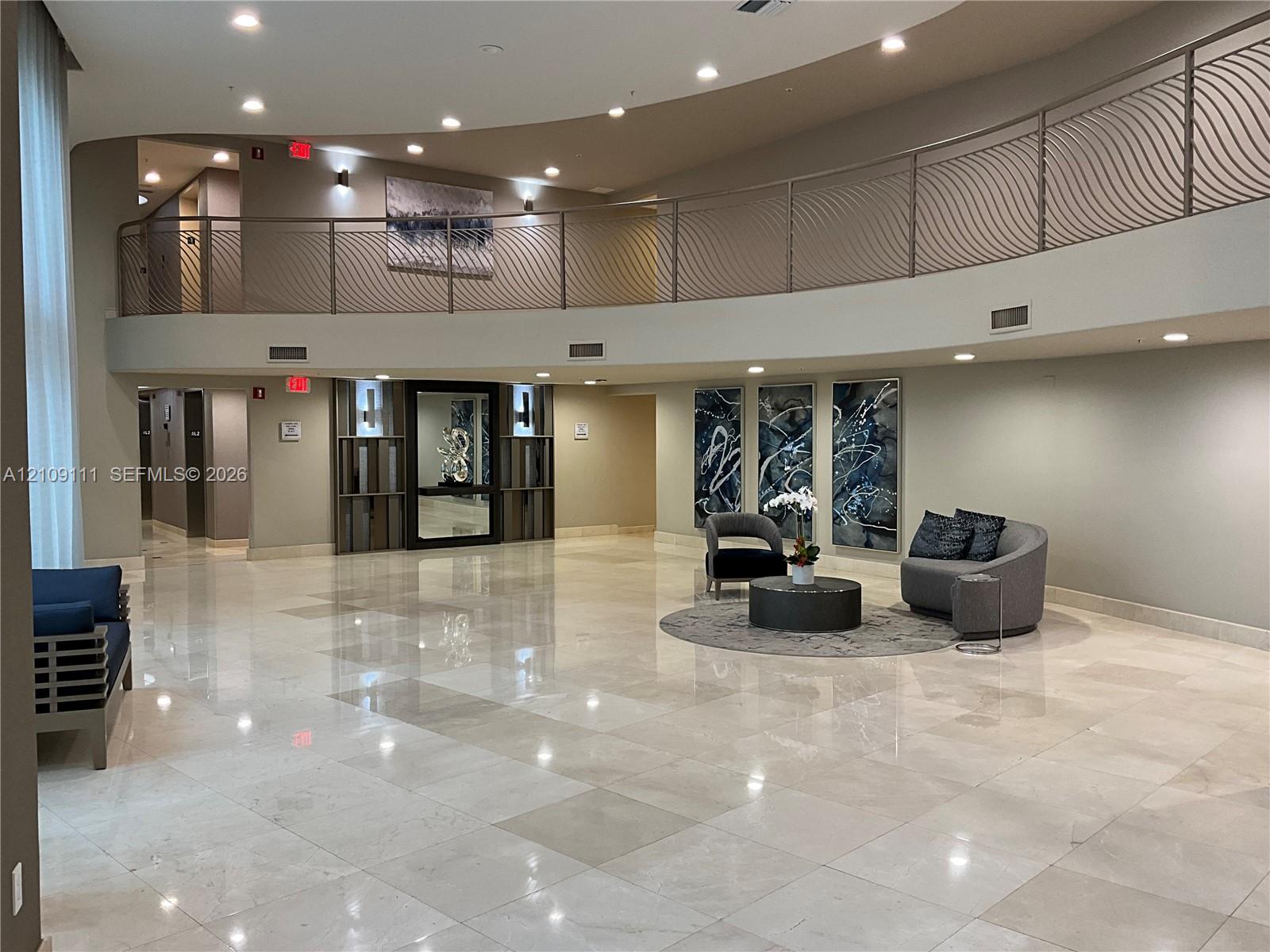 Double-height foyer featuring polished stone flooring, a curved mezzanine with decorative metal railing, recessed ceiling lighting, and built-in wall paneling Double-height foyer featuring polished stone flooring, a curved mezzanine with decorative metal railing, recessed ceiling lighting, and built-in wall paneling