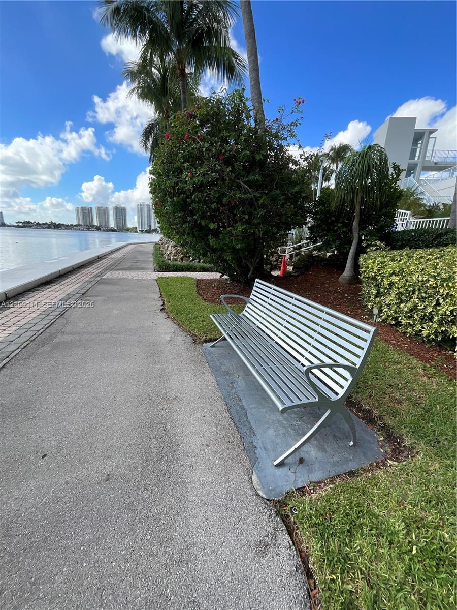 Waterfront promenade featuring a paved walking path, concrete bulkhead, mature palms, and metal outdoor seating