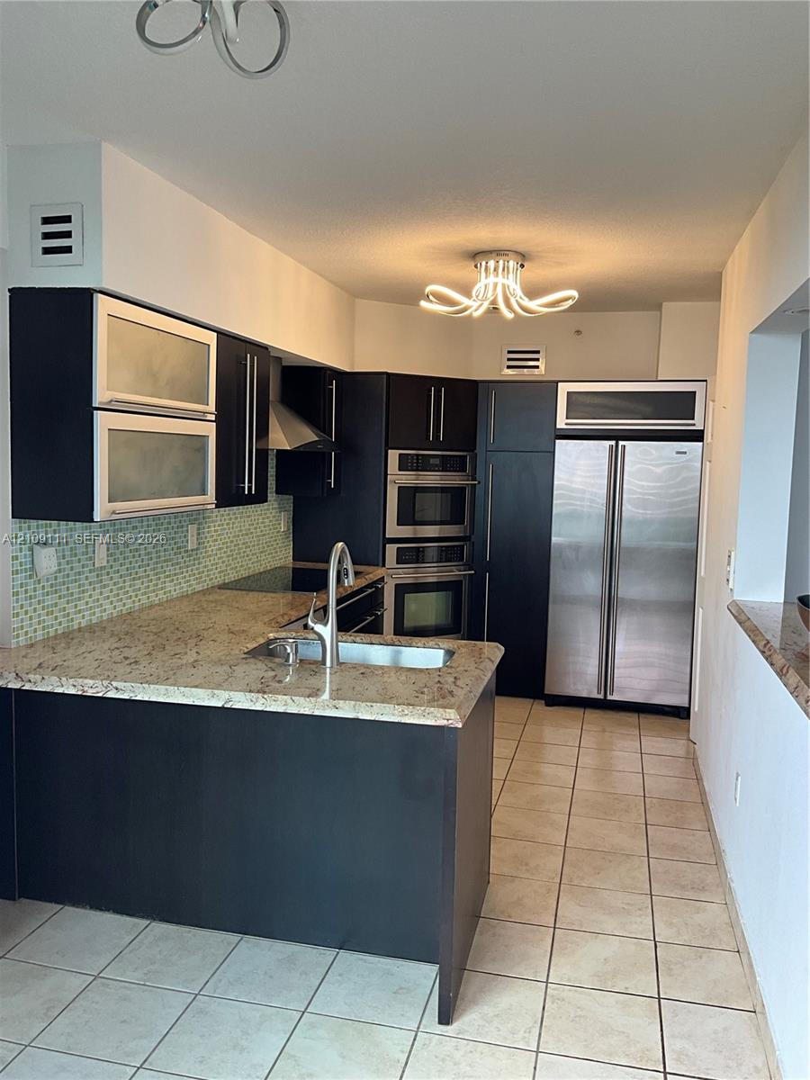 Kitchen featuring dark cabinetry, stone countertops, a mosaic tile backsplash, integrated stainless steel appliances, and a modern tiered chandelier