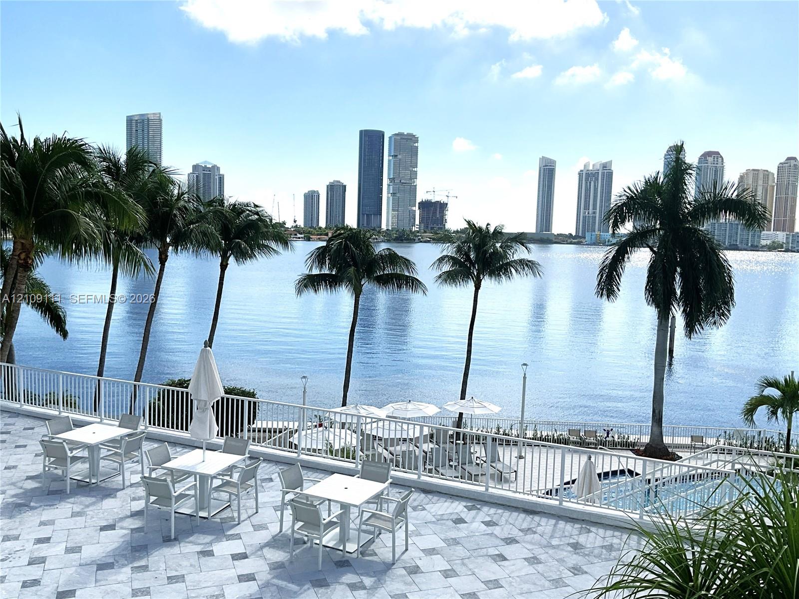 Expansive stone-paved terrace featuring metal railings, an integrated swimming pool, and mature palm trees with a backdrop of waterfront and city skyline Expansive stone-paved terrace featuring metal railings, an integrated swimming pool, and mature palm trees with a backdrop of waterfront and city skyline