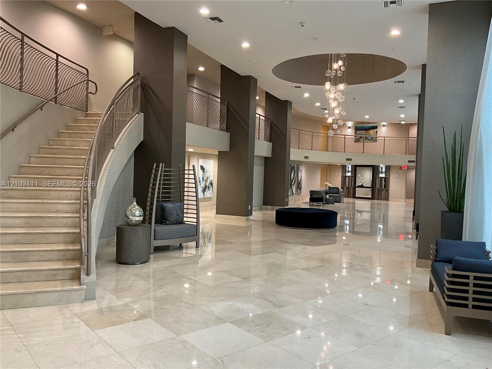 Grand lobby featuring polished stone flooring, a sweeping staircase with custom metal railing, vaulted ceiling with recessed lighting, and a multi-tiered decorative chandelier Grand lobby featuring polished stone flooring, a sweeping staircase with custom metal railing, vaulted ceiling with recessed lighting, and a multi-tiered decorative chandelier
