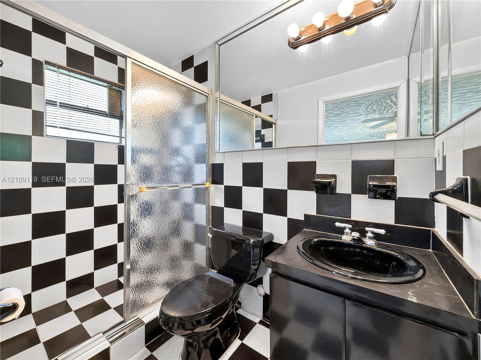 Black and white checkerboard wall and floor tile, glass-enclosed shower, black basin sink with vanity, and large wall-mounted mirror