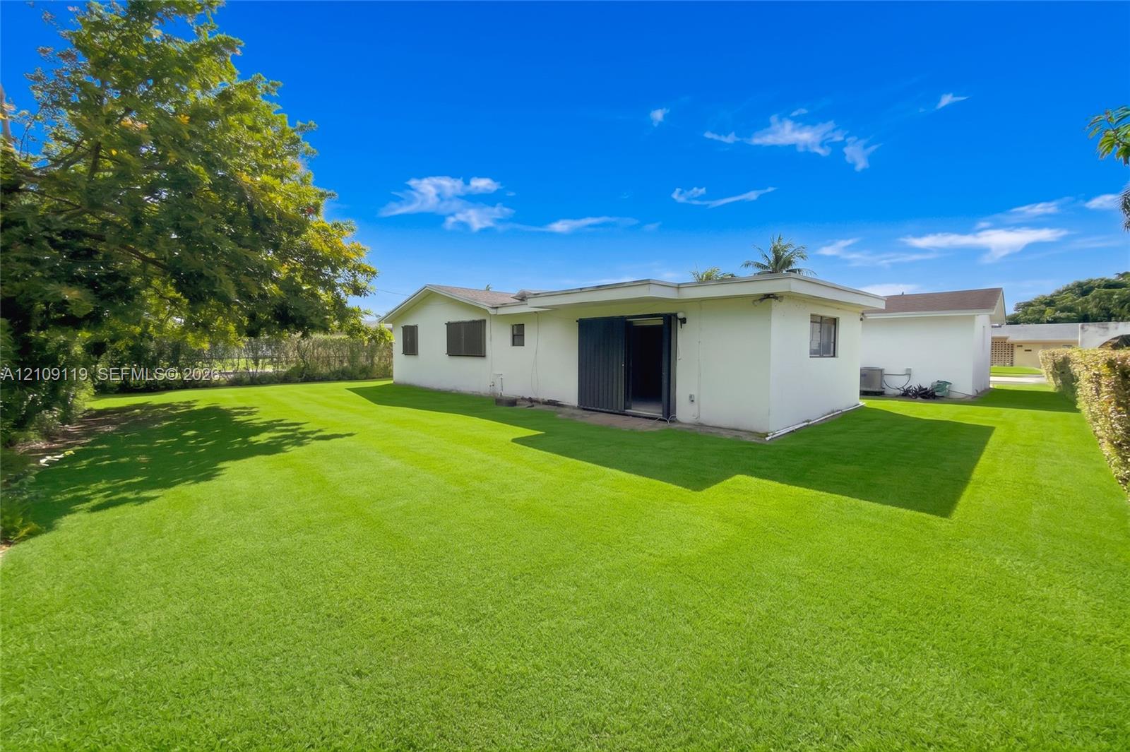 Rear exterior featuring white stucco siding, a flat roofline, sliding glass doors, and an expansive lawn with mature perimeter trees