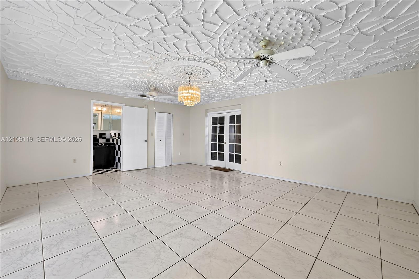 Spacious room featuring a decorative plaster ceiling with medallion accents, large-format tile flooring, a tiered crystal chandelier, a white ceiling fan, and French doors