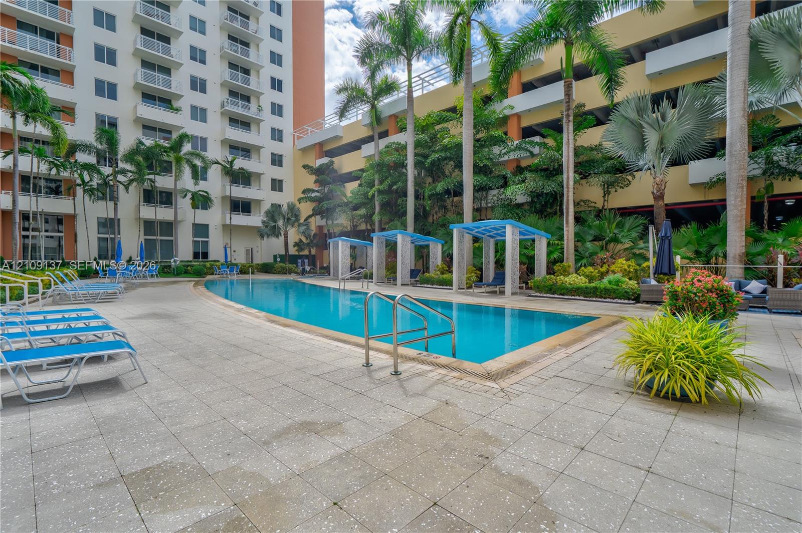 Curved swimming pool with metal handrails, paved pool deck, and shaded pergola structures set among palm trees