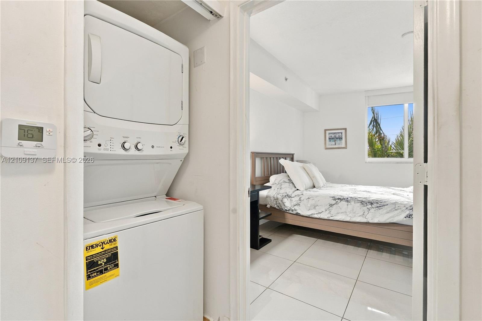 Stackable laundry unit with white finish, large-format tile flooring, and window with exterior foliage