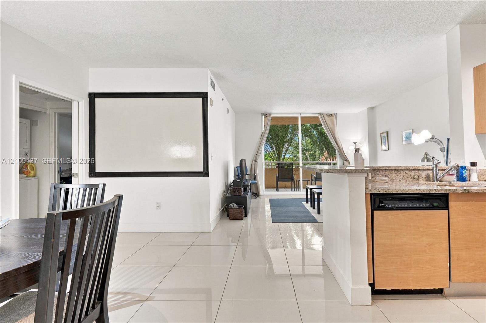 Open-concept interior featuring light-toned tile flooring, kitchen with stone-finish countertops, wood-finish cabinetry, built-in dishwasher, and sliding glass doors to exterior balcony