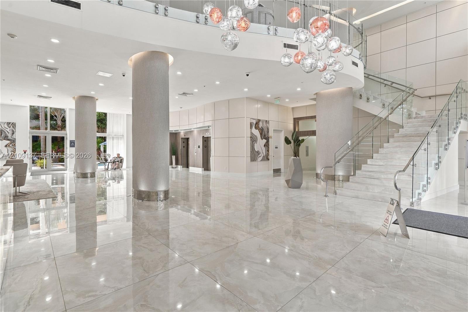 Grand lobby featuring polished stone tile flooring, structural columns with metallic-finish cladding, a multi-globe pendant chandelier, and a glass-paneled staircase