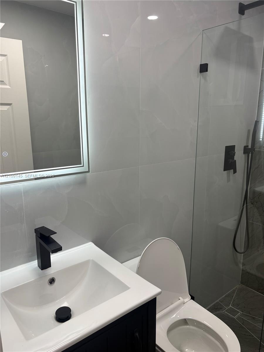 Floating vanity with a white countertop, wall-mounted illuminated mirror, frameless glass shower enclosure, and matte black fixtures