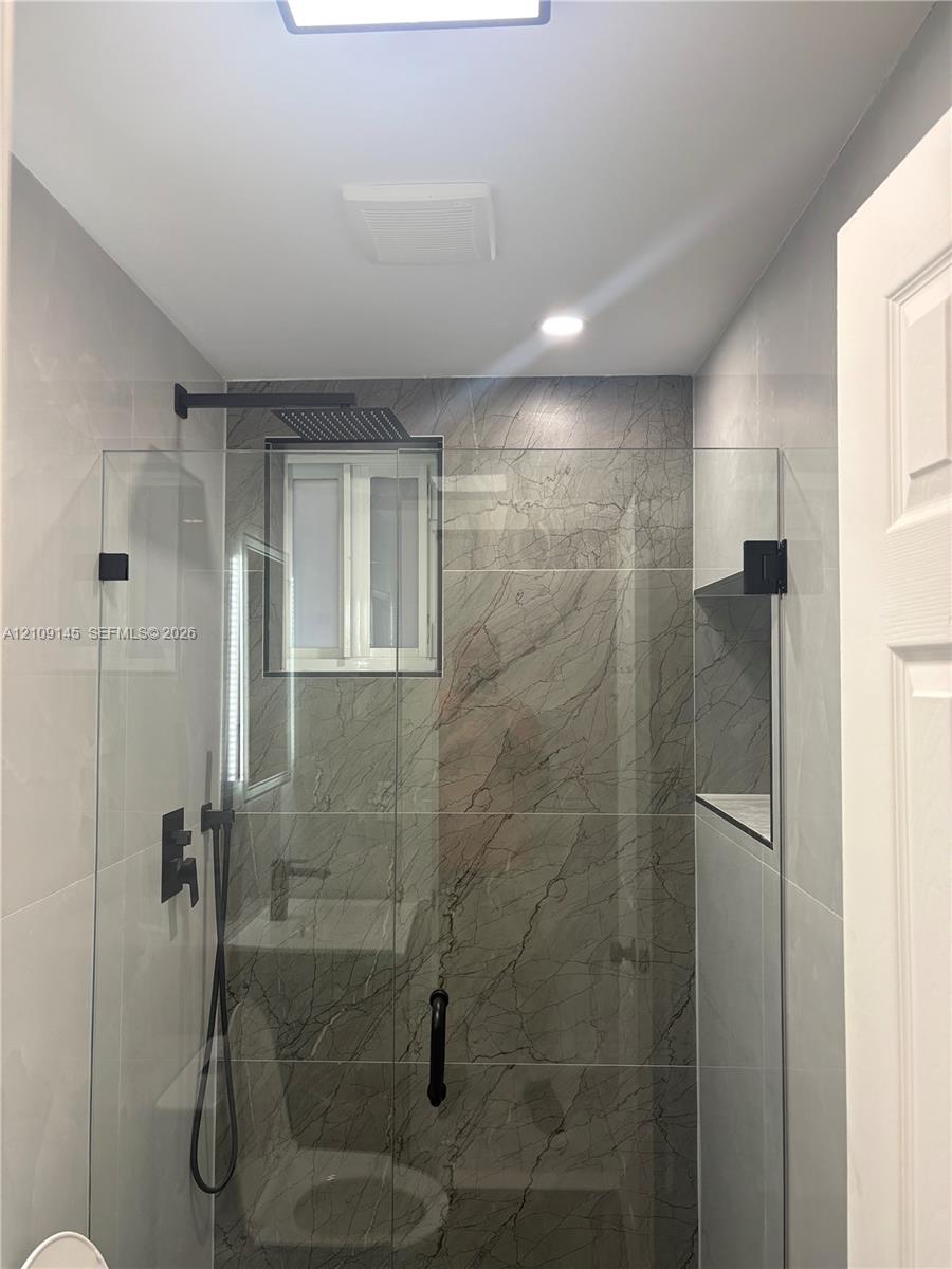 Walk-in shower featuring large-format stone-pattern tile surround, frameless glass enclosure, matte black rainfall shower head, handheld sprayer, and built-in recessed shelf