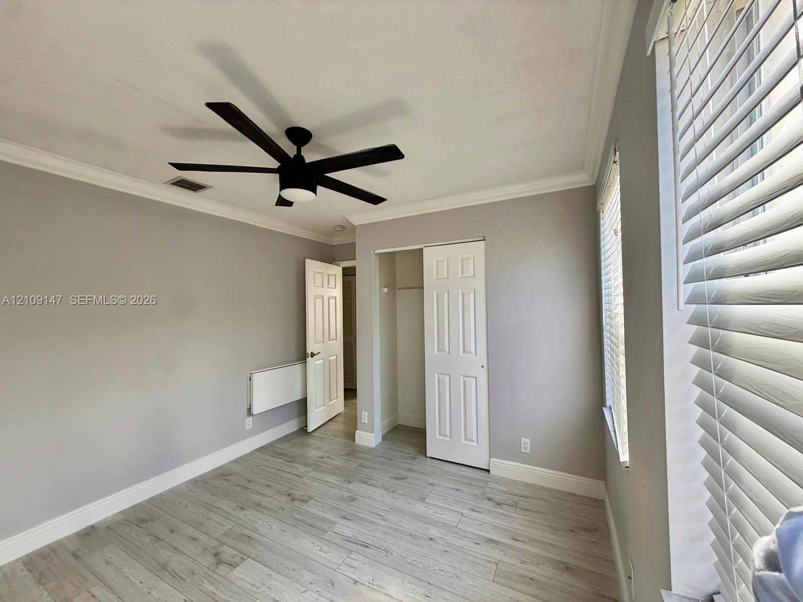 Room featuring wood-finish flooring, crown molding, a multi-blade ceiling fan, and a closet with a six-panel door Room featuring wood-finish flooring, crown molding, a multi-blade ceiling fan, and a closet with a six-panel door
