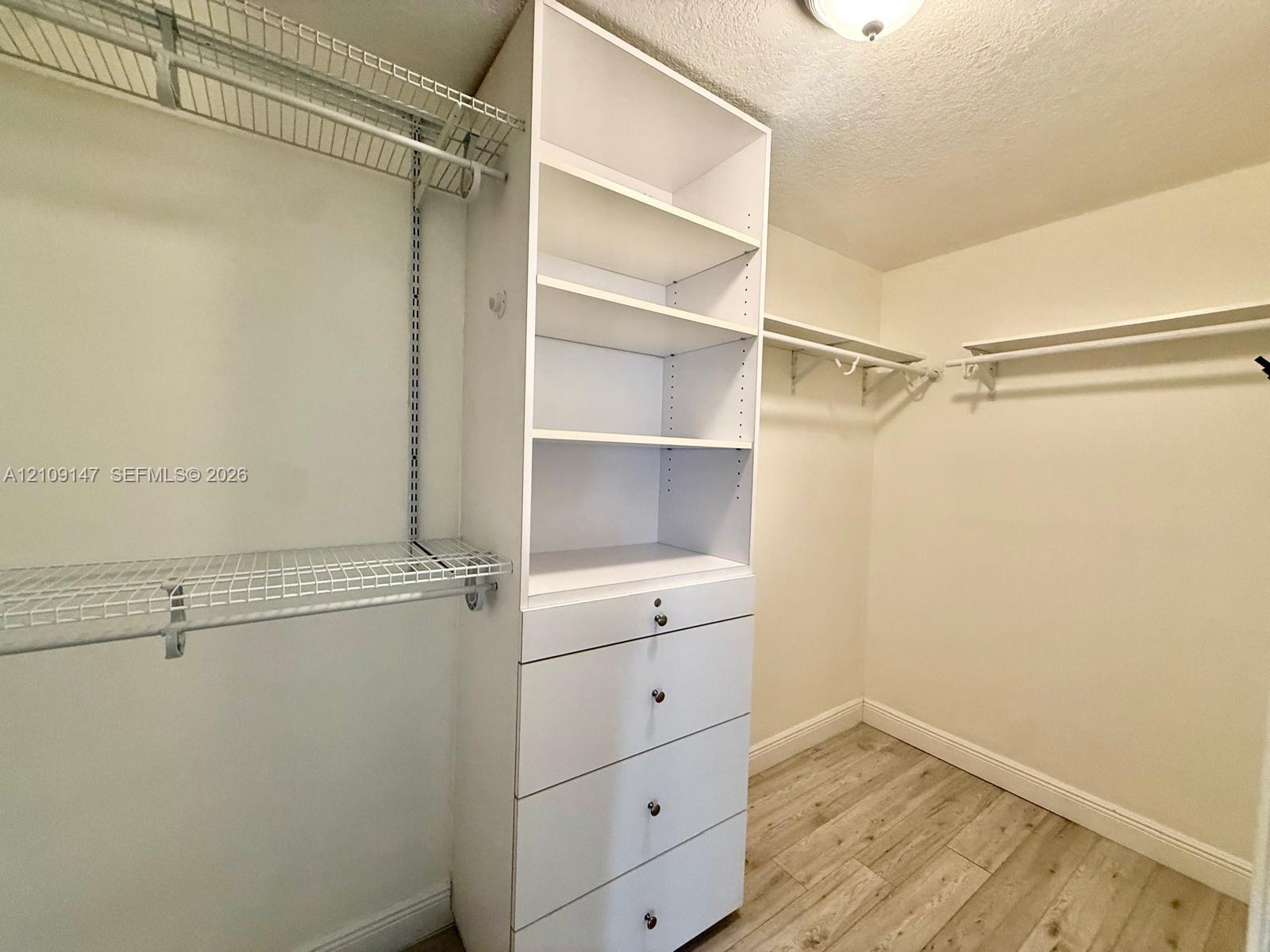 Walk-in closet featuring white built-in shelving, a four-drawer storage unit, metal wire hanging racks, and wood-finish flooring Walk-in closet featuring white built-in shelving, a four-drawer storage unit, metal wire hanging racks, and wood-finish flooring