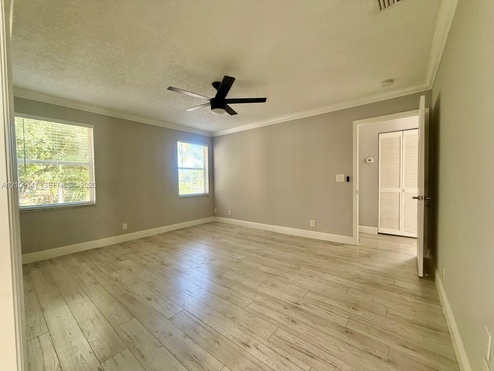 Interior room featuring wood-finish flooring, neutral wall paint, crown molding, a matte black ceiling fan, and louvered-style closet doors Interior room featuring wood-finish flooring, neutral wall paint, crown molding, a matte black ceiling fan, and louvered-style closet doors
