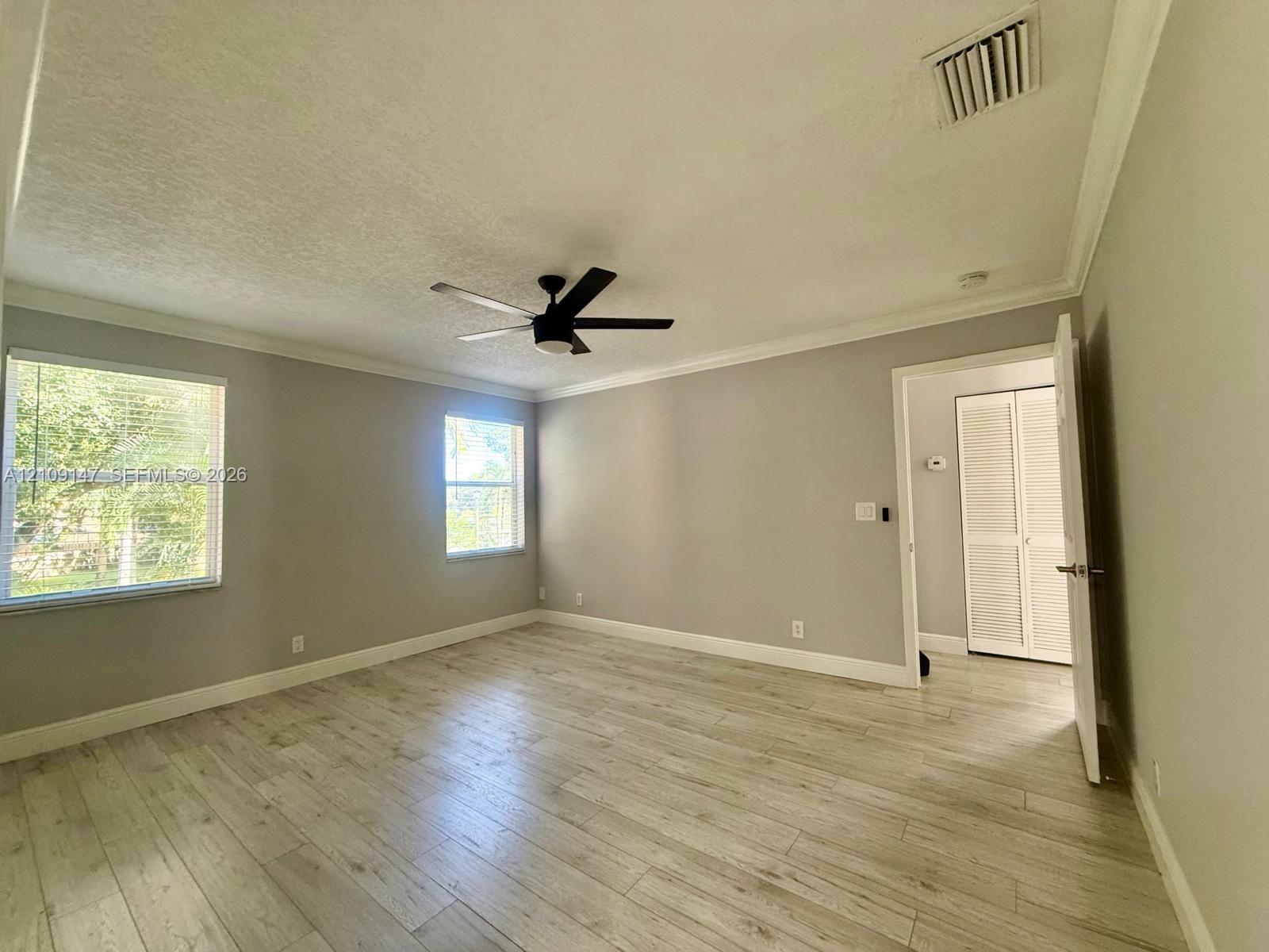 Neutral-toned interior featuring wood-finish flooring, crown molding, multi-pane windows, a ceiling fan, and louvered closet doors Neutral-toned interior featuring wood-finish flooring, crown molding, multi-pane windows, a ceiling fan, and louvered closet doors